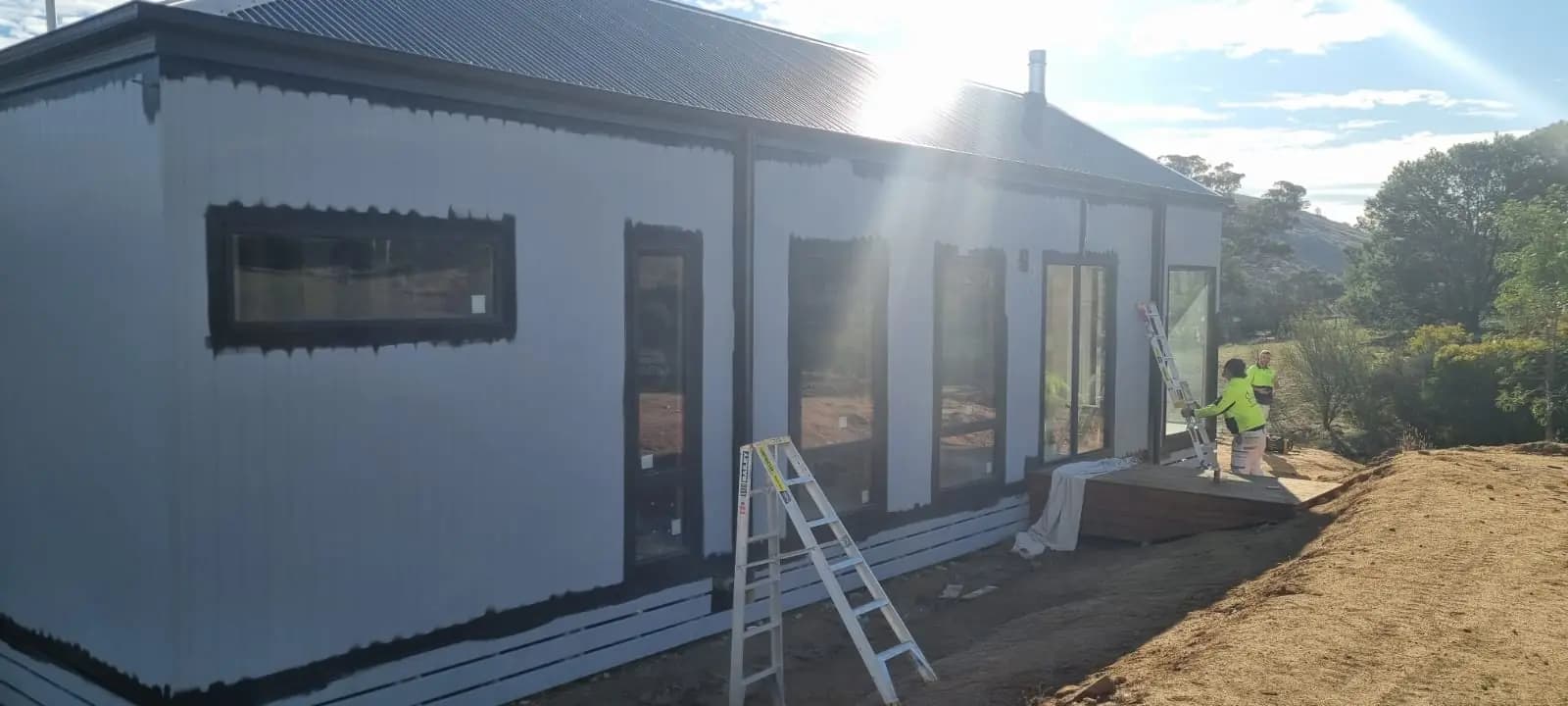 New extension with vertical corrugated cladding in white undercoat, black paint cut in around the window and door frames, a step ladder in front and a worker in a hi-vis shirt at the far end.