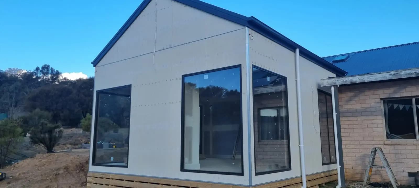 Gable end of a new extension with unpainted pale cladding sheets and large black-framed corner windows, joined to an existing cream brick house with a blue roof.