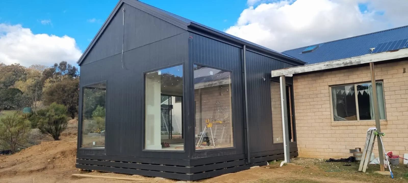 Gable end of a new extension finished in charcoal, with large black-framed corner windows, standing beside an existing cream brick house.