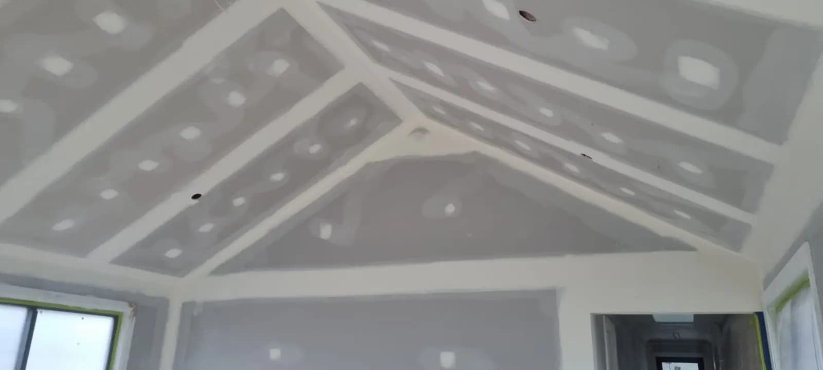 Raked plasterboard ceiling with the joints set and the whole surface in undercoat, masking tape around the window heads and downlight cut-outs visible.