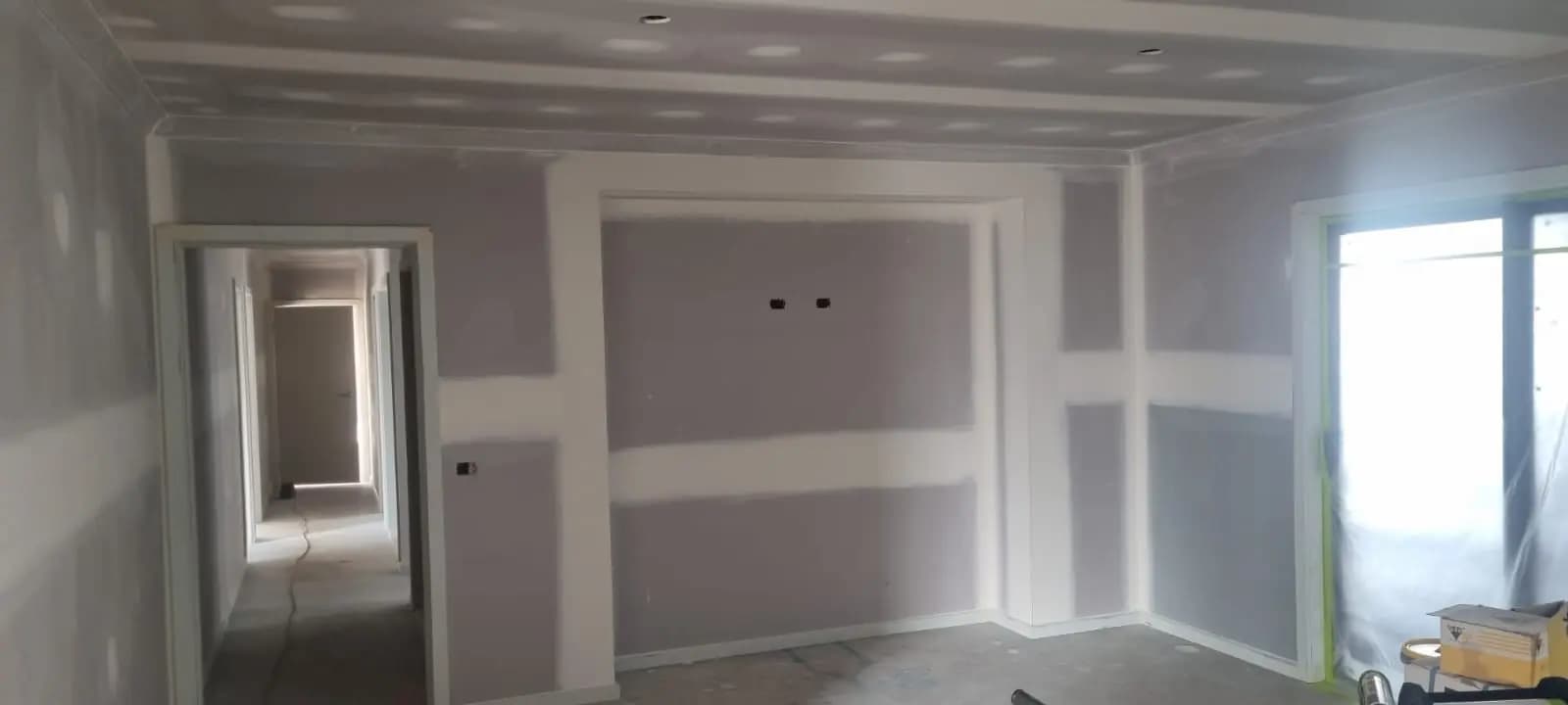 Living area of a new house with bare plasterboard walls and ceiling, set joints still showing, a hallway beyond the doorway and a masked sliding door on the right.