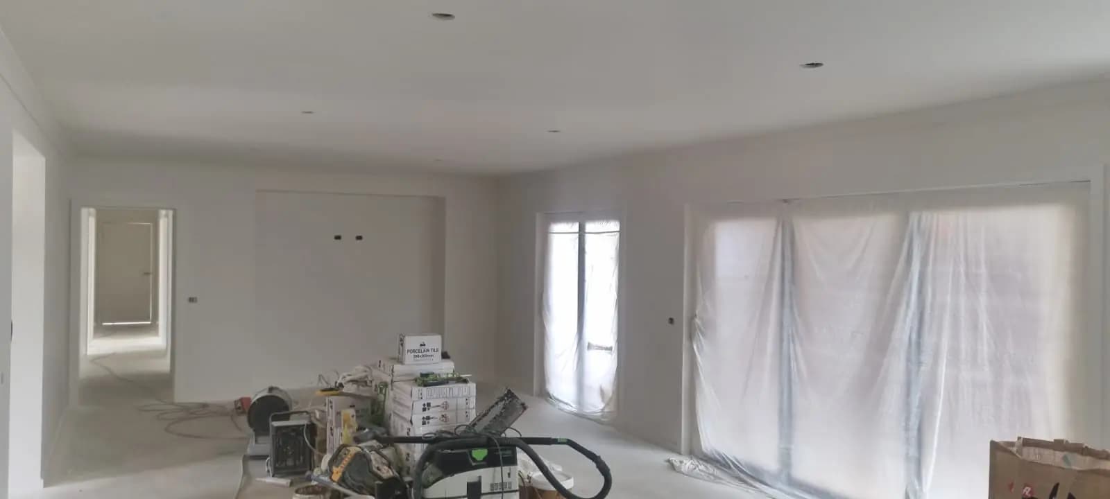 Large open-plan room in a new house with walls and ceiling painted white, windows and a door masked with plastic, and boxes of tiles and power tools on the floor.