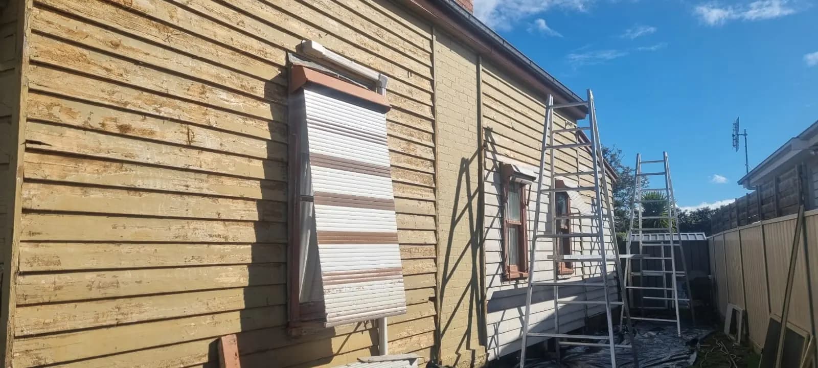 Close view along a weatherboard wall stripped back to bare timber, with a sheet of corrugated iron propped over a window opening and ladders set up further along the wall.