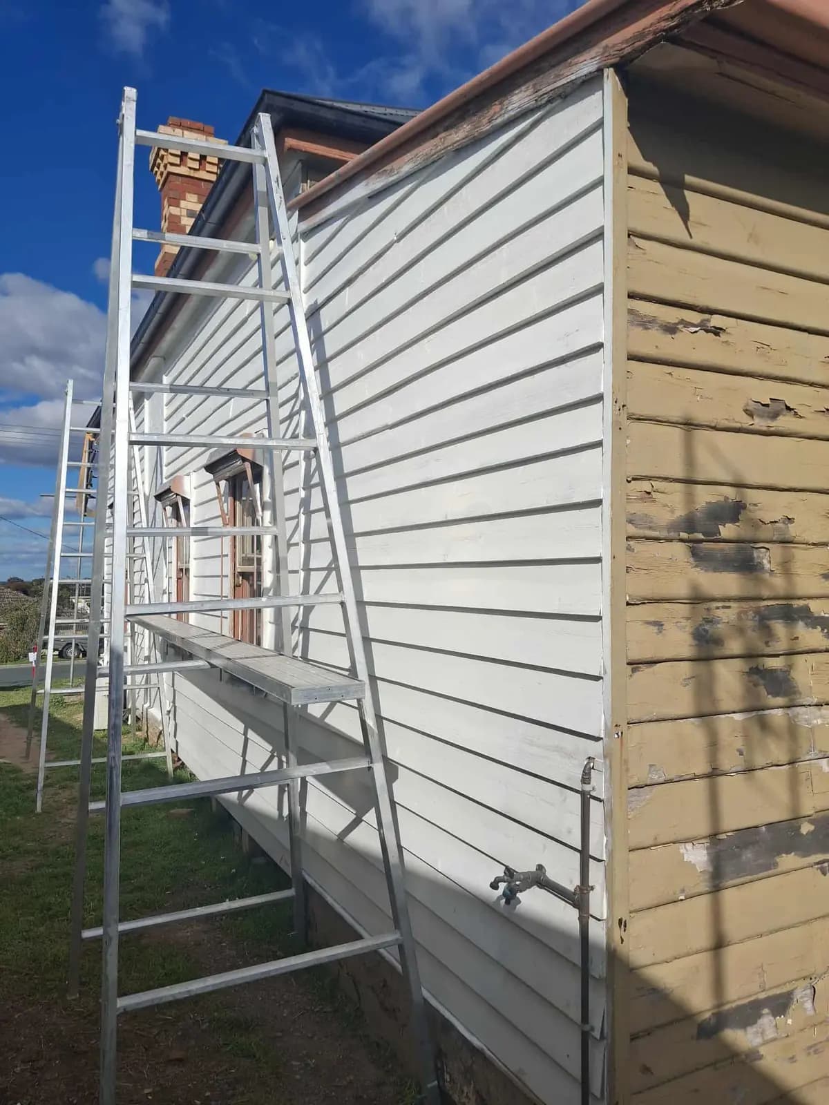 Corner of a weatherboard cottage showing freshly painted white boards next to a section of old cream paint that is cracked and flaking, with scaffold planks in front.