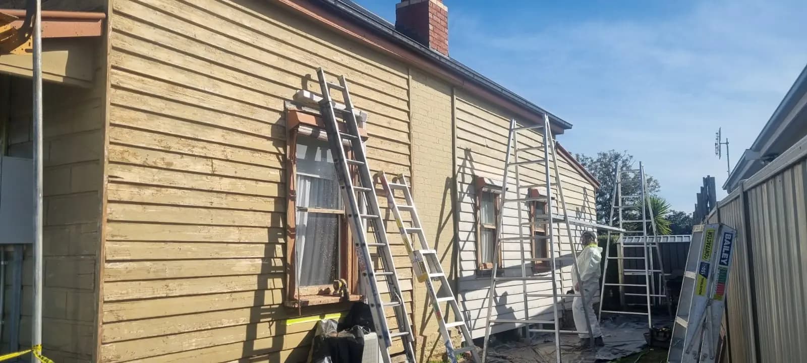Side wall of a weatherboard cottage with the old paint stripped back to bare timber, aluminium ladders and trestles leaning against the wall and a painter in white overalls working near the red brick chimney.