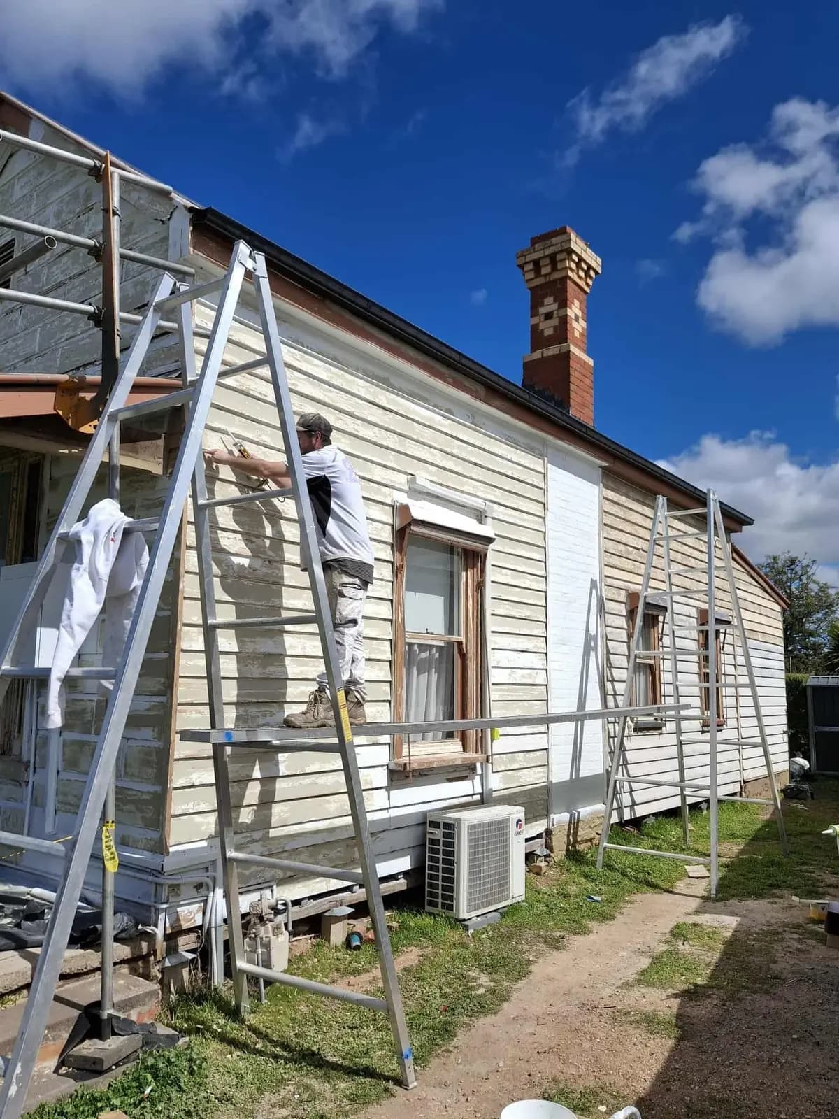 A painter standing on a trestle plank sanding a weatherboard wall that is part way through being painted white, with a red brick chimney and a split-system unit below.